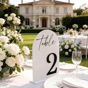 May include: A white arched acrylic table number sign featuring the black script text Table and a large black numeral 2. The sign is positioned on a white tablecloth next to a glass, a white plate, and a bouquet of white roses in a clear vase.