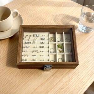K&ouml;nnte beinhalten: Eine Holzschmuckschatulle mit Glasdeckel, gef&uuml;llt mit verschiedenen Ohrringen und Ringen. Die Ohrringe sind silberfarben und umfassen Stecker, Anh&auml;nger und Creolen. Die Ringe sind ebenfalls silberfarben und in separaten F&auml;chern ausgestellt.
