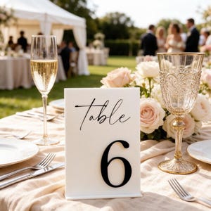 May include: A white rectangular table number sign with the word Table in black script and the number 6 in a large black serif font. The sign is placed on a table set with a tall champagne flute, a decorative glass goblet, and silverware.
