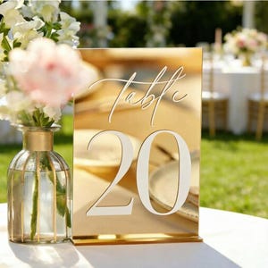 May include: A rectangular gold mirror table sign featuring the white text "Table 20" in a combination of elegant script and bold serif fonts. The sign has a reflective surface and stands upright on a flat base, displaying a clear number twenty.