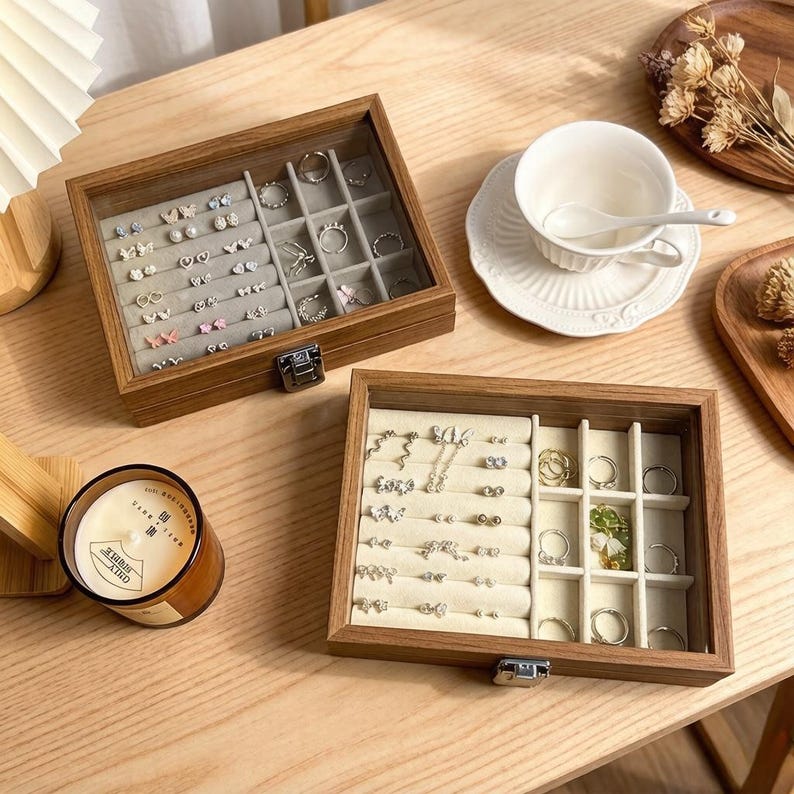 K&ouml;nnte beinhalten: Zwei Schmuckk&auml;stchen aus Holz mit Glasdeckeln, gef&uuml;llt mit Ohrringen und Ringen. Die Boxen sind ge&ouml;ffnet und zeigen die organisierten F&auml;cher. Eine brennende Kerze und eine Teetasse mit Untertasse befinden sich ebenfalls auf der Holzoberfl&auml;che.