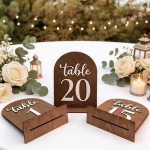 Könnte beinhalten: Tischkarten aus Holz mit dunkelbraunem Finish. Die Schilder zeigen das Wort "Tisch" in Schreibschrift und große weiße Zahlen. Die Tischnummern 1, 15 und 20 sind sichtbar. Ideal für Hochzeiten oder Veranstaltungen.