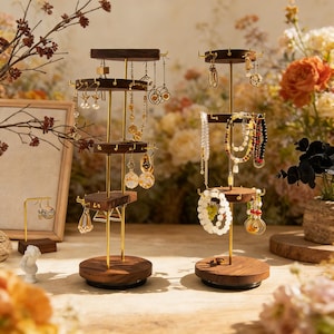 May include: Two wooden jewellery display stands with multiple tiers and gold-coloured metal supports. The stands hold various earrings, necklaces, and bracelets. The bases are round and dark brown. The background features flowers and a framed picture.