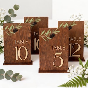 Pode incluir: Sinais de números de mesa de madeira com design de ramo de oliveira. Os sinais são castanhos com letras douradas e apresentam os números 5, 10 e 12. O design do ramo de oliveira inclui folhas verdes e azeitonas pretas. Ideal para casamentos ou eventos.