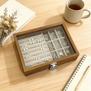 K&ouml;nnte beinhalten: Eine Holzschmuckschatulle mit Glasdeckel, gef&uuml;llt mit verschiedenen Ohrringen und Ringen. Die Ohrringe haben Schmetterlings-, Perlen- und Herzmotive. Die Ringe sind silberfarben und haben unterschiedliche Stile. Die Schatulle steht auf einer Holzoberfl&auml;che.