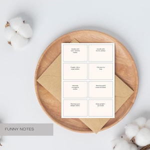 Könnte beinhalten: Ein Satz von acht cremefarbenen Karten mit humorvollen Phrasen, präsentiert auf einem Holztablett mit einem braunen Umschlag. Der Text enthält Sätze wie "Handle with care - dreams inside" und "This box is a hug."