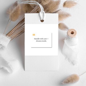 Pu&ograve; includere: Un'etichetta regalo bianca con un cordino, con il testo "Handle with care&mdash;dreams inside." L'etichetta &egrave; ambientata su uno sfondo di fiori secchi, nastro bianco e una bobina di legno, creando un'estetica minimalista.