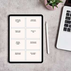 Pu&ograve; includere: Un tablet nero mostra otto riquadri bianchi con testo, tra cui "Handle with care - dreams inside" e "This box is a hug". Uno stilo bianco e un laptop sono nelle vicinanze, insieme a una piccola pianta in vaso. Il tablet &egrave; su una superficie grigia.