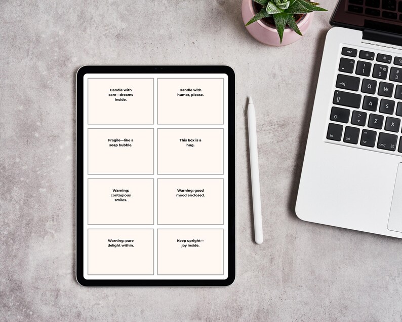 Pu&ograve; includere: Un tablet nero mostra otto riquadri bianchi con testo, tra cui "Handle with care - dreams inside" e "This box is a hug". Uno stilo bianco e un laptop sono nelle vicinanze, insieme a una piccola pianta in vaso. Il tablet &egrave; su una superficie grigia.