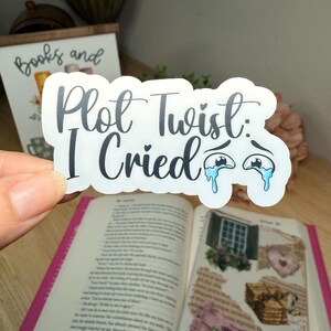 Puede incluir: Una pegatina blanca con el texto "Plot Twist: I Cried" en escritura negra, con ojos de dibujos animados y lágrimas azules. La pegatina se sostiene sobre un libro abierto con una cubierta rosa.