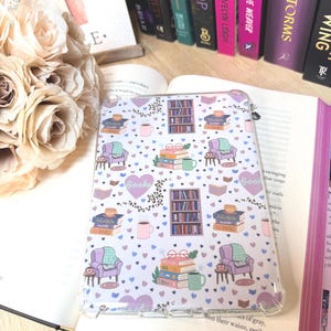 Puede incluir: Funda transparente para teléfono con un patrón repetido de libros, sillones, tazas de café y estanterías. El diseño incluye la palabra "books" en forma de corazón. La funda está sobre un libro abierto con otros libros en el fondo.