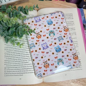 Könnte beinhalten: Eine transparente Tablet-Hülle mit einem skurrilen Muster aus Eulen, Sonnenblumen und roten Schleifen auf weißem Hintergrund. Die Hülle liegt auf einem offenen Buch, mit einer grünen Pflanze und Büchern im Hintergrund.