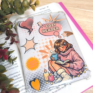 May include: Clear phone case with a comic-style illustration of a woman reading with a mug, candles, and the text "COZY READER." The design features hearts, stars, and speech bubbles in orange, pink, and yellow.