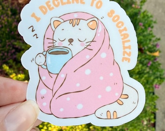 Könnte beinhalten: Weißer Aufkleber mit einer Cartoon-Katze, die in eine rosa Decke mit weißen Punkten gehüllt ist und eine blaue Tasse hält. Der Aufkleber trägt den Text "I DECLINE TO SOCIALIZE" in Orange.