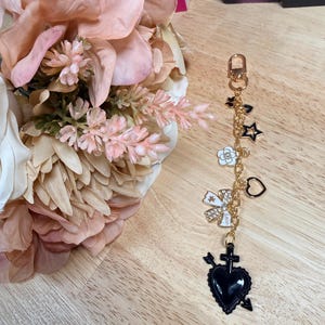 May include: A gold-toned keychain with various charms, including a black heart pierced by an arrow, a star, a bow, and a flower. The charms are connected by a delicate gold chain. The background includes a bouquet of pink flowers.