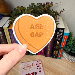 May include: An orange heart-shaped sticker with the text "AGE GAP". The sticker has a glittery effect and is held in front of a backdrop of books and greenery. A Valentine's Day card with hearts and a gift box is visible.