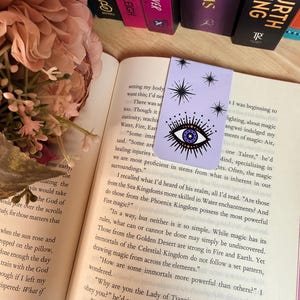May include: A lavender bookmark with a celestial eye design, featuring an eye with a blue iris and stars. The bookmark is placed in an open book with text. Books with titles like "STORMS" and "FOURTH WING" are visible in the background.