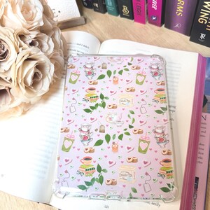 May include: Transparent tablet case with a pink design of teacups, hearts, tea bags, and biscuits. The case rests on an open book, with a backdrop of cream roses and a row of books. The text "ENGLISH TEA" is printed on the case.