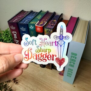 May include: A holographic sticker featuring a line-art illustration of a dagger piercing a red heart. The text reads Soft Heart Sharp Dagger in a decorative font. The sticker has a reflective, rainbow-colored finish that shifts in the light.