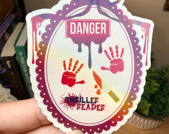 Könnte beinhalten: Aufkleber mit einem lila-weißen, gewellten Rand, mit einer Schleife oben und dem Wort "DANGER" in einem roten Rechteck. Rote Handabdrücke, ein Messer und die Worte "THRILLER READER" sind ebenfalls vorhanden.