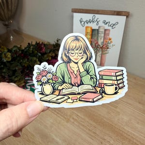 May include: A sticker featuring a cartoon illustration of a person reading at a desk. The person wears glasses and a green cardigan. Books, a coffee cup, and a vase of flowers are on the desk. A sign behind the person reads "Books and".