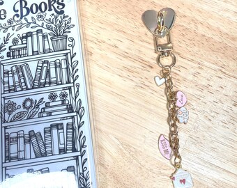 Könnte beinhalten: Transparente Handyhülle mit einem schwarz-weißen Bücherregal-Design und dem Wort "Books". Ein goldfarbener Schlüsselanhänger mit Herz- und Charm-Akzenten, darunter ein weißer Umschlag, ein rosa Herz und ein "Kiss Me"-Anhänger.