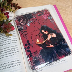 May include: A clear phone case featuring a gothic illustration of a woman reading a book with a candle. The case has a red background with black roses and vines. The phone case is on top of an open book with text.
