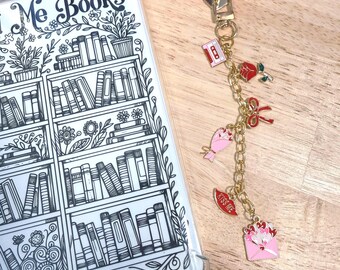 Könnte beinhalten: Eine transparente Handyhülle mit einem schwarz-weißen Bücherregal-Design und dem Text "Me Books". Ein goldener Charm mit Herzverschluss zeigt verschiedene Charms, darunter eine Kassette, eine Rose und einen "Kiss Me" Lippen-Charm.