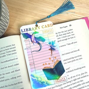 May include: A whimsical bookmark featuring a library card design with a blue tassel. The bookmark showcases illustrations of a dragon, fairy, and an open book. The text "LIBRARY CARD" and the number "031211" are visible.