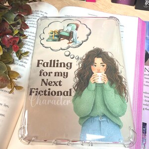 Puede incluir: Funda transparente para teléfono con una ilustración impresa de una mujer con un suéter verde y jeans, sosteniendo una taza. El texto "Falling for my Next Fictional Character" y una burbuja con una silla y libros.