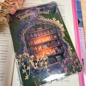 Può includere: Una custodia protettiva trasparente con un design a tema fantasy. L'opera d'arte raffigura una libreria con candele, libri e bottiglie di pozioni, incorniciata da un arco e da un fogliame. Le parole "Fantasy Forest" sono in alto.