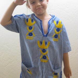 May include: A light blue tunic-style shirt with short sleeves and a V-neck. The shirt features yellow and blue embroidered geometric designs on the front and pockets. Blue trim accents the neckline, sleeves, and hem. Two pockets are visible.