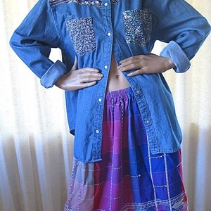 May include: A blue denim shirt with floral embroidery on the pockets and yoke. The shirt is worn over a multi-colored skirt with fringe trim. The skirt has a plaid pattern with pink, blue, and white stripes.