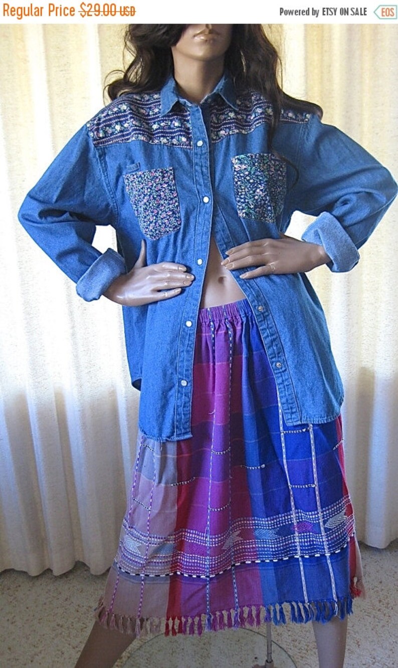 May include: A blue denim shirt with floral embroidery on the pockets and yoke. The shirt is worn over a multi-colored skirt with fringe trim. The skirt has a plaid pattern with pink, blue, and white stripes.