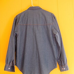 May include: A blue denim shirt with long sleeves and a button-down collar. The shirt is hanging on a white hanger against a yellow wall.