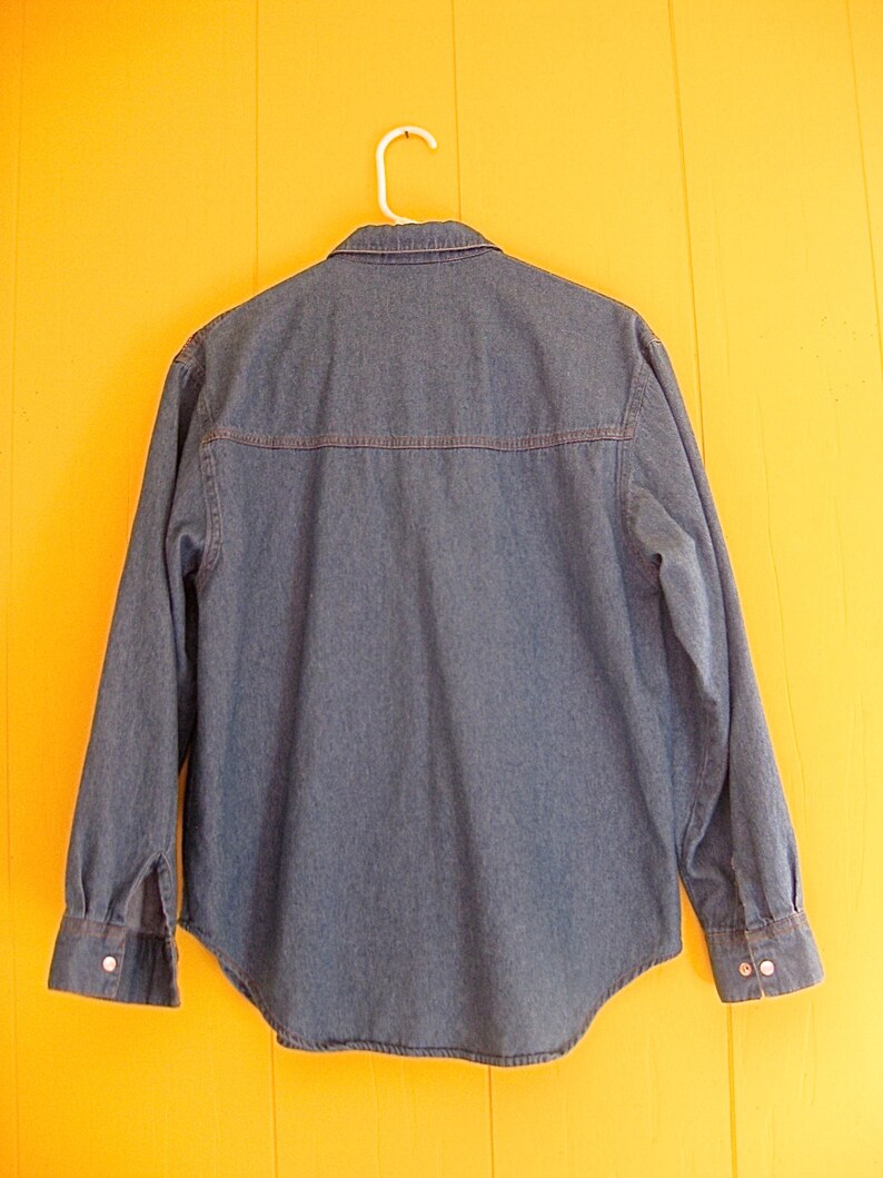 May include: A blue denim shirt with long sleeves and a button-down collar. The shirt is hanging on a white hanger against a yellow wall.