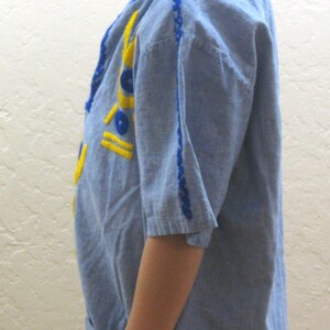 May include: A light blue tunic-style shirt with yellow and blue embroidered details. The shirt has short sleeves and a pocket. The model is wearing a blue headband with a Superman logo.