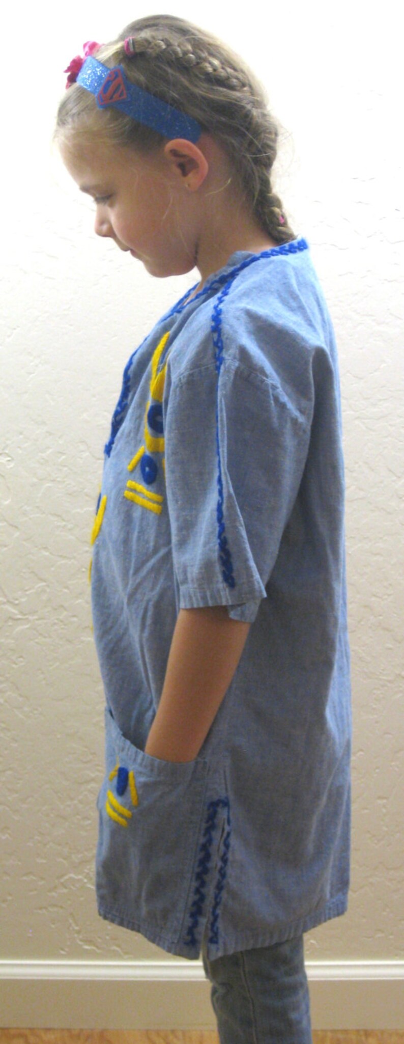 May include: A light blue tunic-style shirt with yellow and blue embroidered details. The shirt has short sleeves and a pocket. The model is wearing a blue headband with a Superman logo.