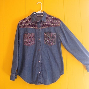 May include: A blue denim shirt with floral patterned fabric accents on the collar and pockets. The shirt has a button-up front and long sleeves.