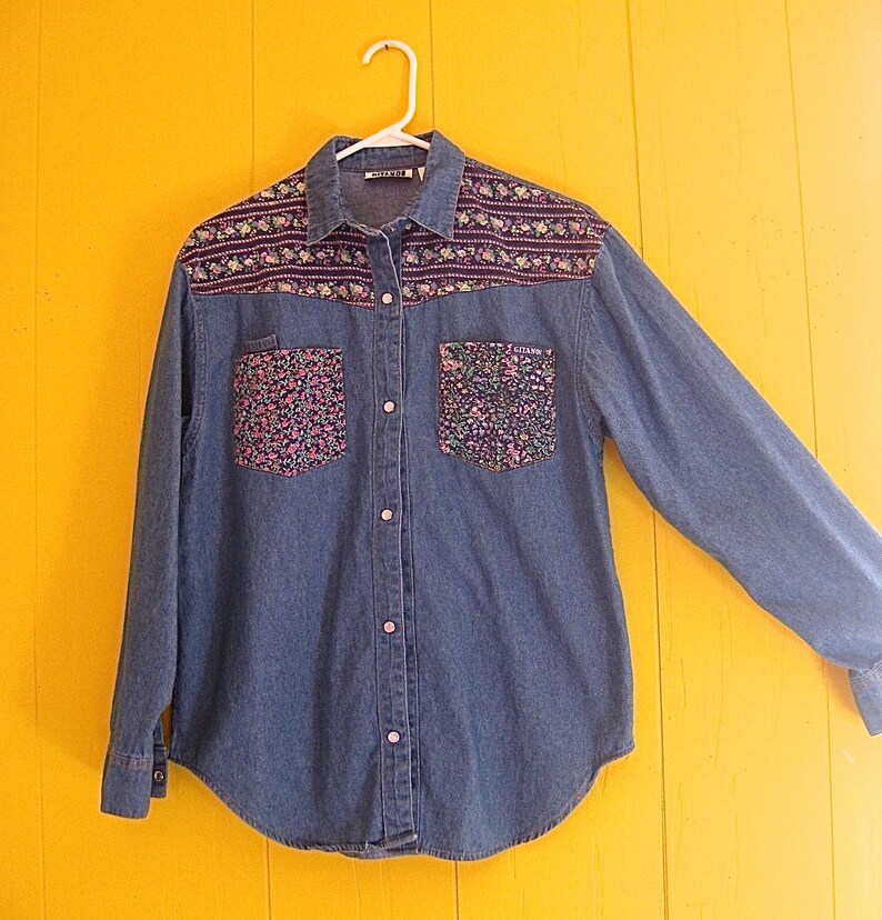 May include: A blue denim shirt with floral patterned fabric accents on the collar and pockets. The shirt has a button-up front and long sleeves.