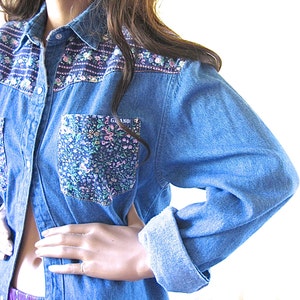 May include: A blue denim shirt with a floral print pocket and floral trim around the collar. The shirt has a button-up front and long sleeves.