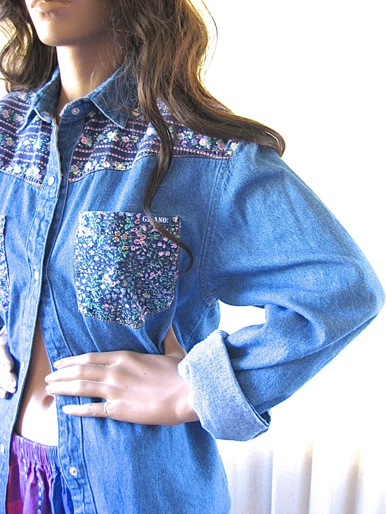 May include: A blue denim shirt with a floral print pocket and floral trim around the collar. The shirt has a button-up front and long sleeves.