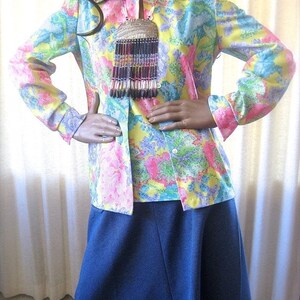 May include: A vintage floral print blouse with a long-sleeved button-up design. The blouse is yellow, pink, and blue with a multi-colored beaded necklace. The model is wearing a dark blue skirt.