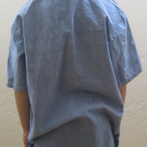 May include: A light blue linen tunic with short sleeves and a blue embroidered neckline. The tunic is paired with light blue denim jeans. The subject has blonde hair styled in braids.