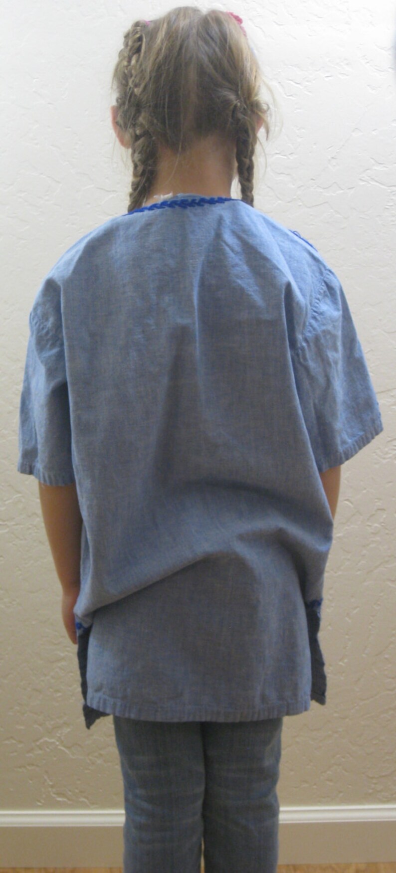 May include: A light blue linen tunic with short sleeves and a blue embroidered neckline. The tunic is paired with light blue denim jeans. The subject has blonde hair styled in braids.