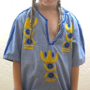 May include: A light blue tunic-style shirt with yellow and blue embroidered details. The shirt has short sleeves, a v-neck, and two front pockets. The neckline and sleeves are trimmed with blue stitching. A young person is wearing the shirt.