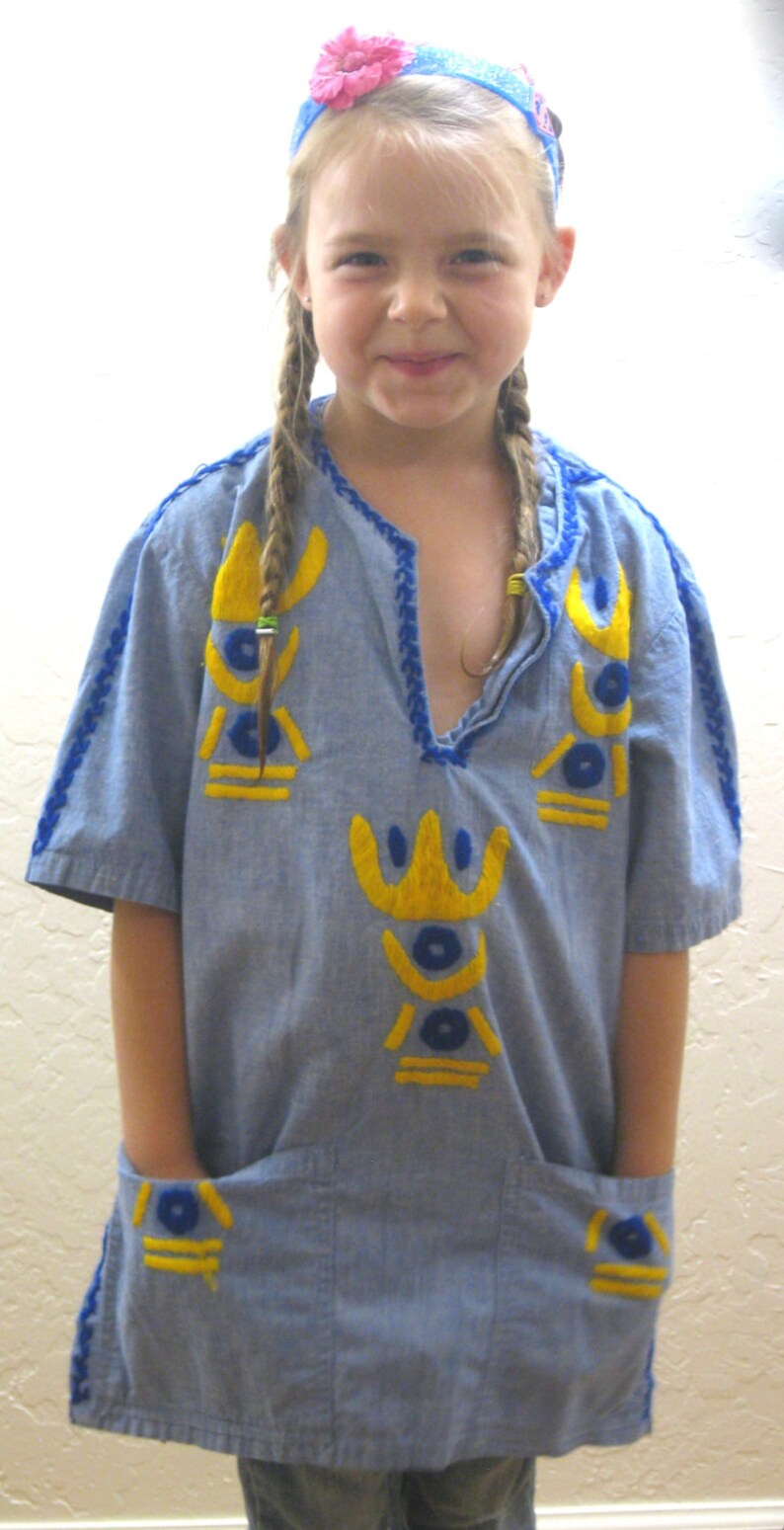 May include: A light blue tunic-style shirt with yellow and blue embroidered details. The shirt has short sleeves, a v-neck, and two front pockets. The neckline and sleeves are trimmed with blue stitching. A young person is wearing the shirt.