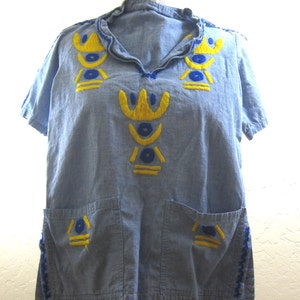 May include: A light blue short-sleeved shirt with yellow and blue embroidered geometric designs. The shirt has two front pockets and a ruffled collar. The edges of the sleeves and the collar are trimmed with blue stitching.