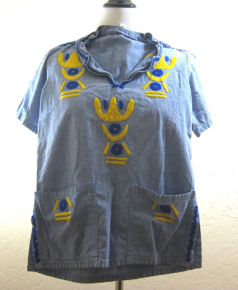 May include: A light blue short-sleeved shirt with yellow and blue embroidered geometric designs. The shirt has two front pockets and a ruffled collar. The edges of the sleeves and the collar are trimmed with blue stitching.