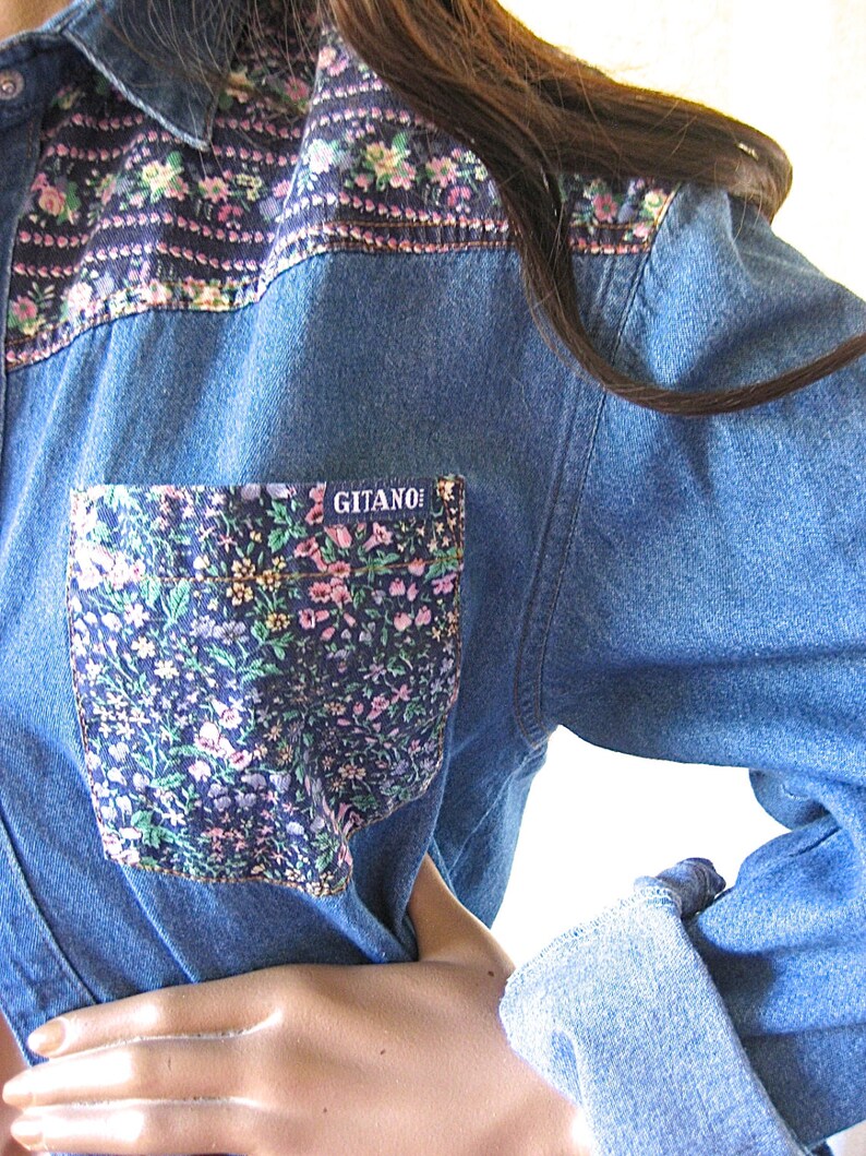 May include: A blue denim shirt with a floral print on the collar and a pocket. The pocket has a floral print and the word "GITANO" in white letters.